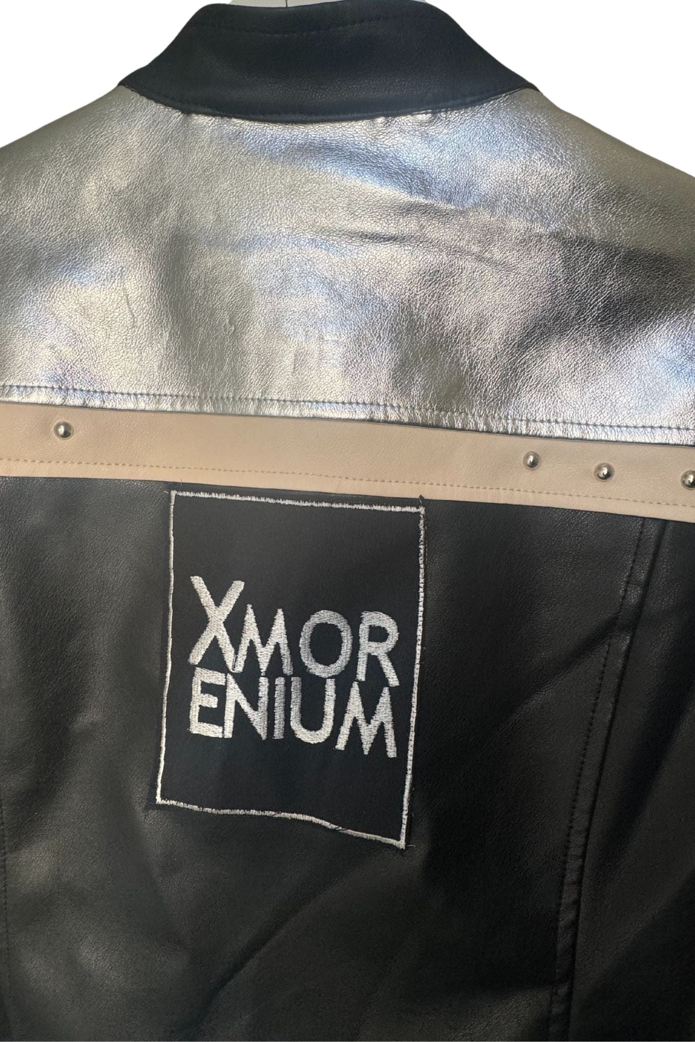 Rückseite der silber-schwarzen Kunstlederjacke mit großem Xmorenium-Logo und beiger Zierleiste mit Nieten.