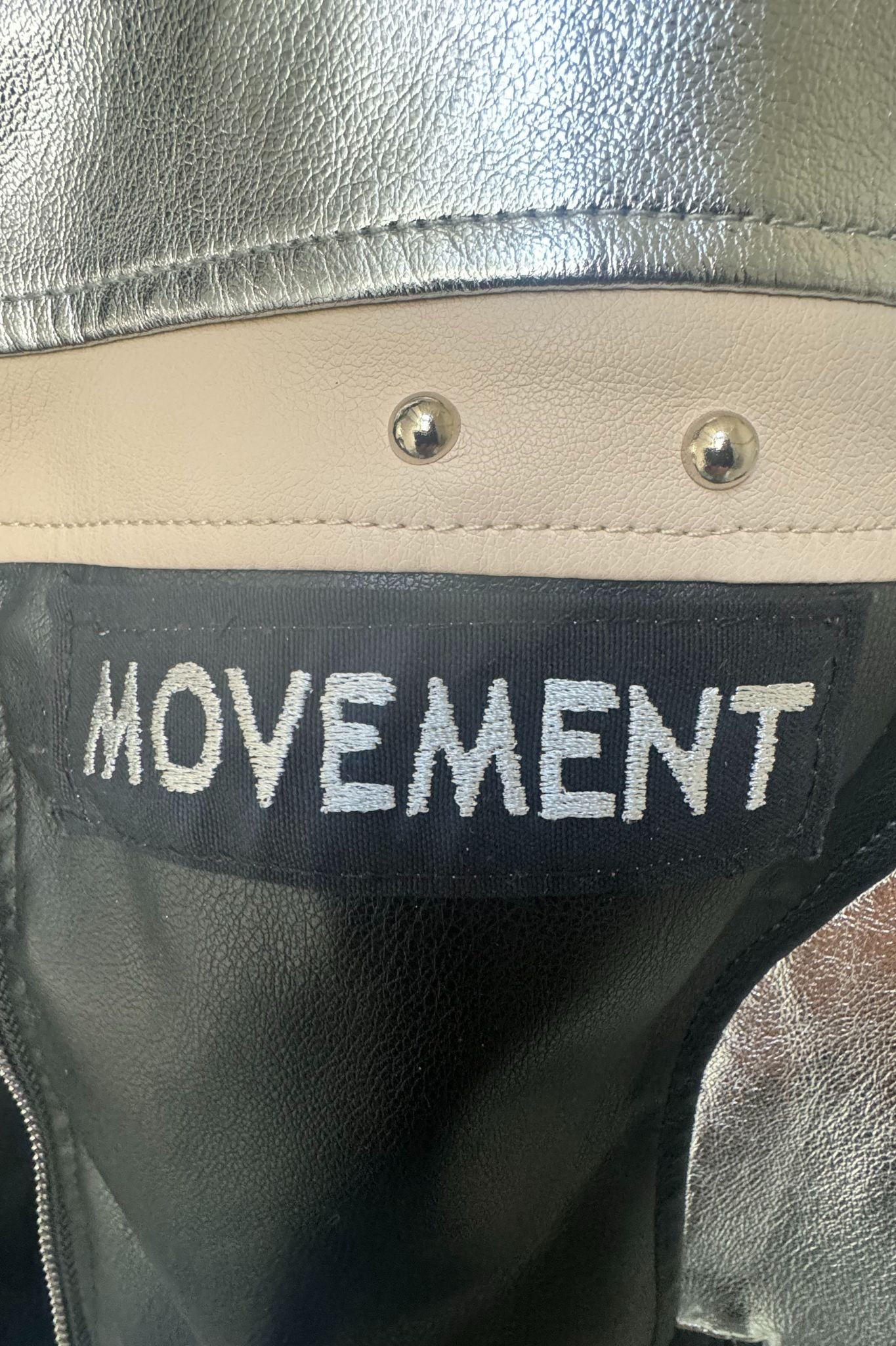 Detailaufnahme des Movement-Schriftzugs auf der silber-schwarzen Kunstlederjacke mit beiger Ziernaht und Nieten.