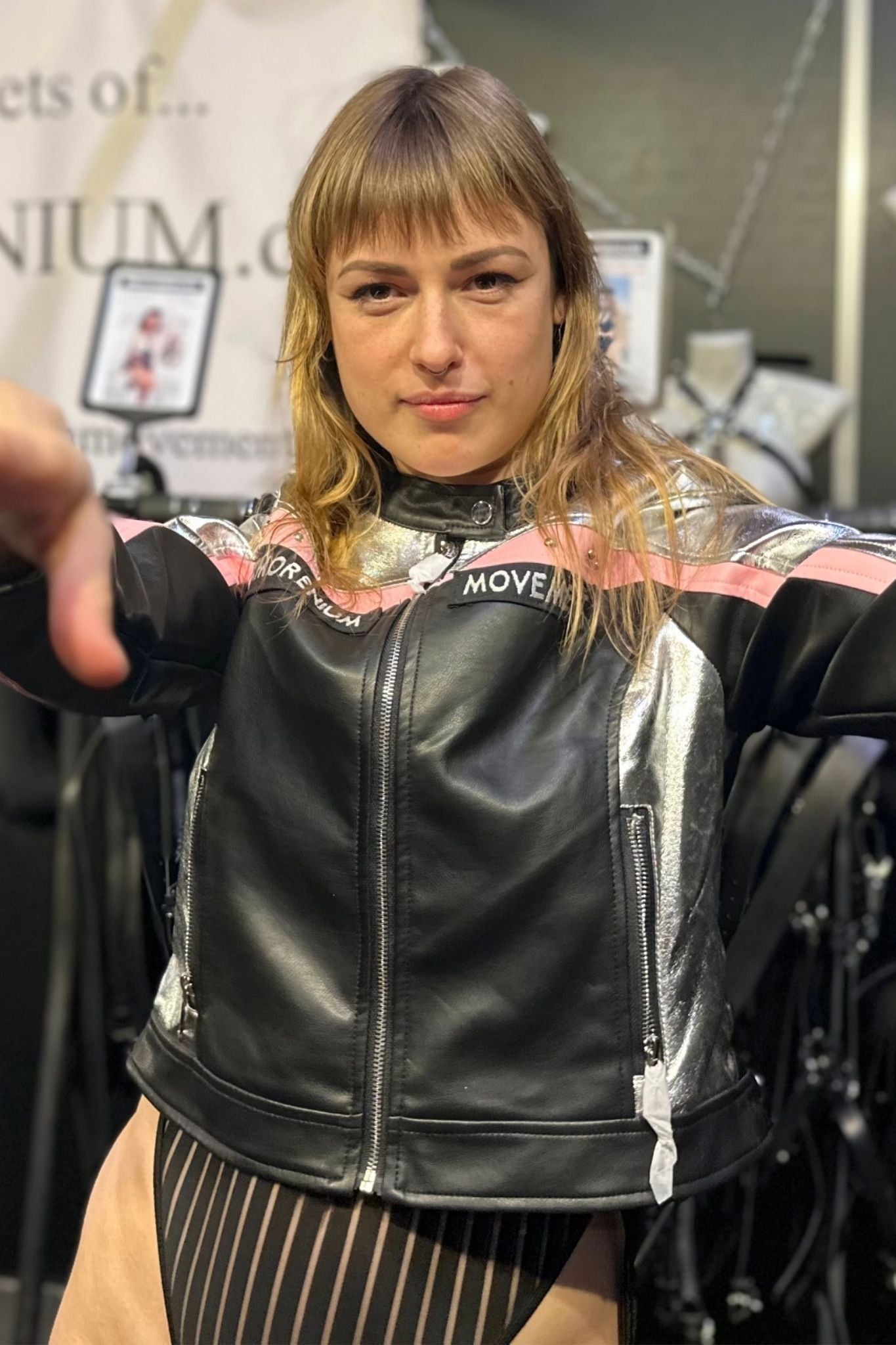 Frau trägt silber-schwarze Kunstlederjacke mit rosa Akzenten und Xmorenium-Patch auf Messe, zeigt selbstbewusste Pose Richtung Kamera.